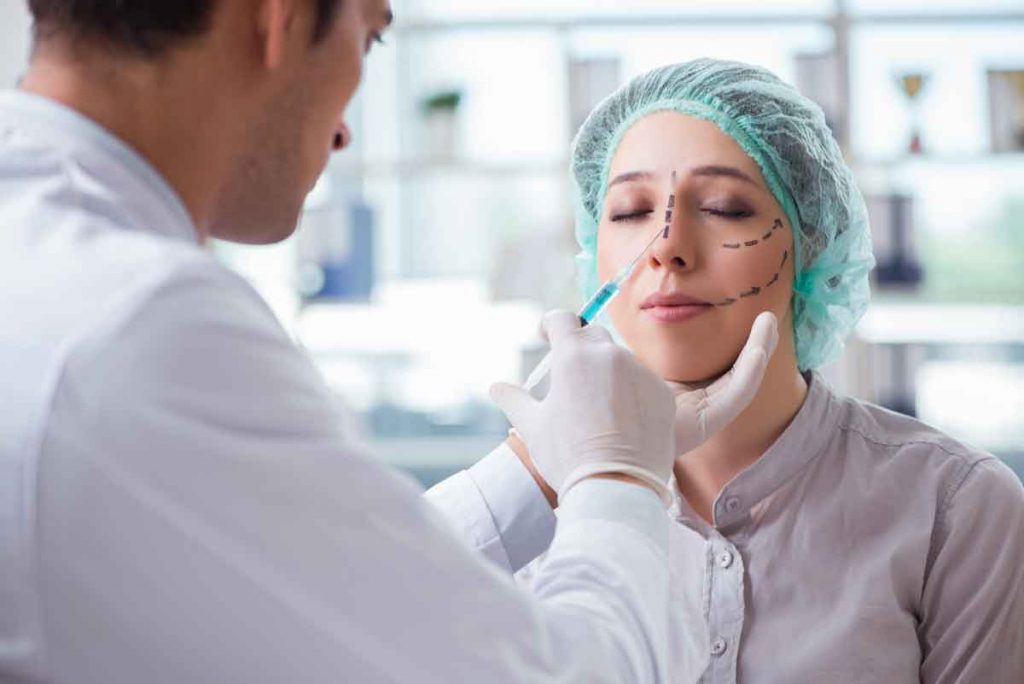 Woman with bouffant hat/hair net on, with lines for surgery drawn on face, with doctor/nurse about to inject blue liquid into nose