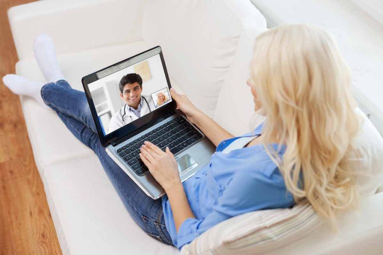 Blonde woman sitting on sofa having telehealth appointment with doctor on laptop