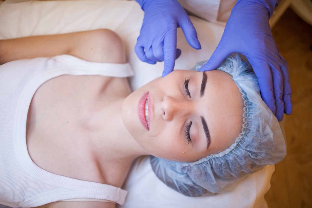 Woman eyes closed, with hair in bouffant hat laying on table with gloved nurse touching face