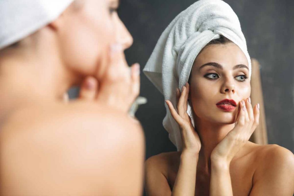 Reflection in mirror of woman with hair up in towel with fingers touching cheeks looking off to the side