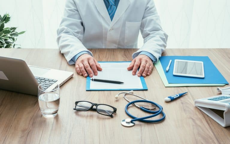 Dr. sitting at desk with water, glasses, stethoscope, thermometer, paper, pen, laptop, pen, notebook and file folders on desk