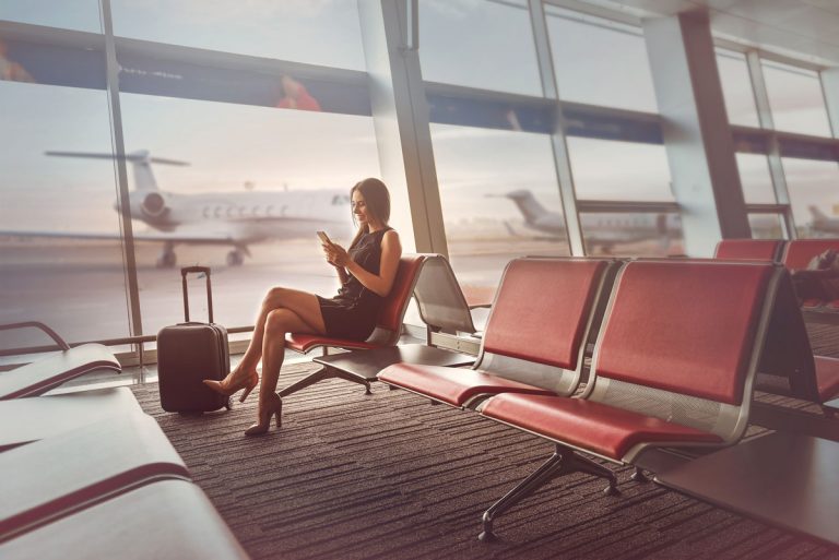 Woman in dress with carry on luggage sitting by window and waiting in airport terminal