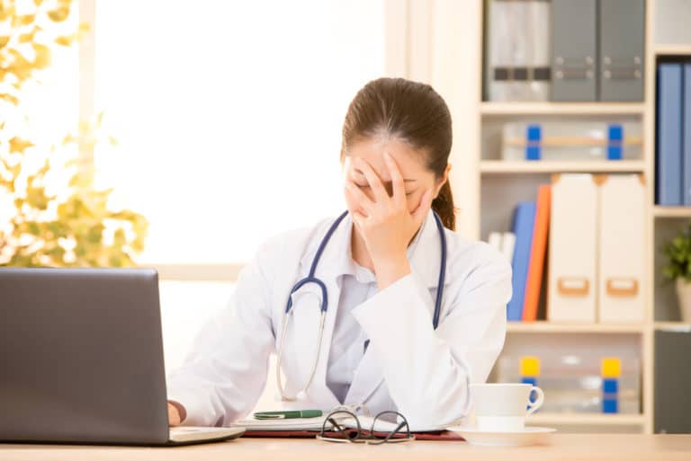 Worried practitioner wearing stethoscope with her hand covering her face sitting in front of lap top