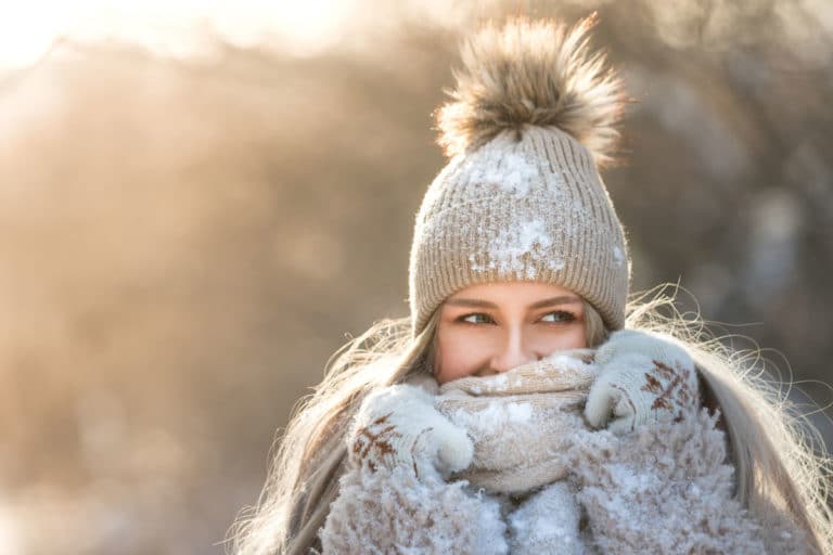 Young woman with hat, coat, scarf and gloves with snow on them smiling and looking side eyed