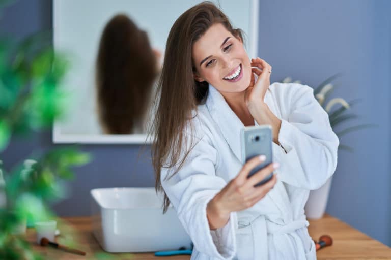 Woman in robe smiling at her cell phone with mirror behind her
