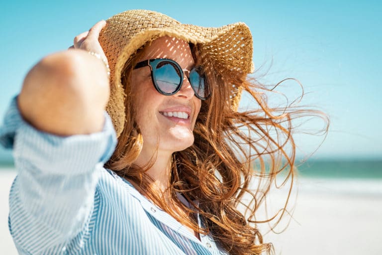 Smiling woman at the beach in a wide brimmed hat, sunglasses and long sleeved shirt