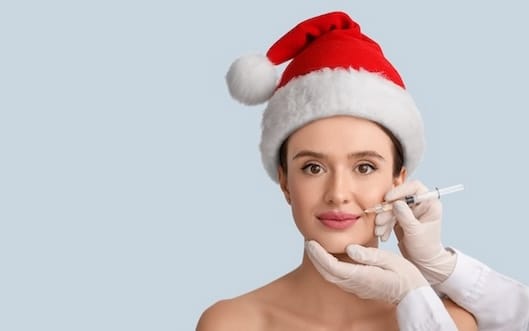 Young woman in Santa hat receiving filler injection in lips against light background